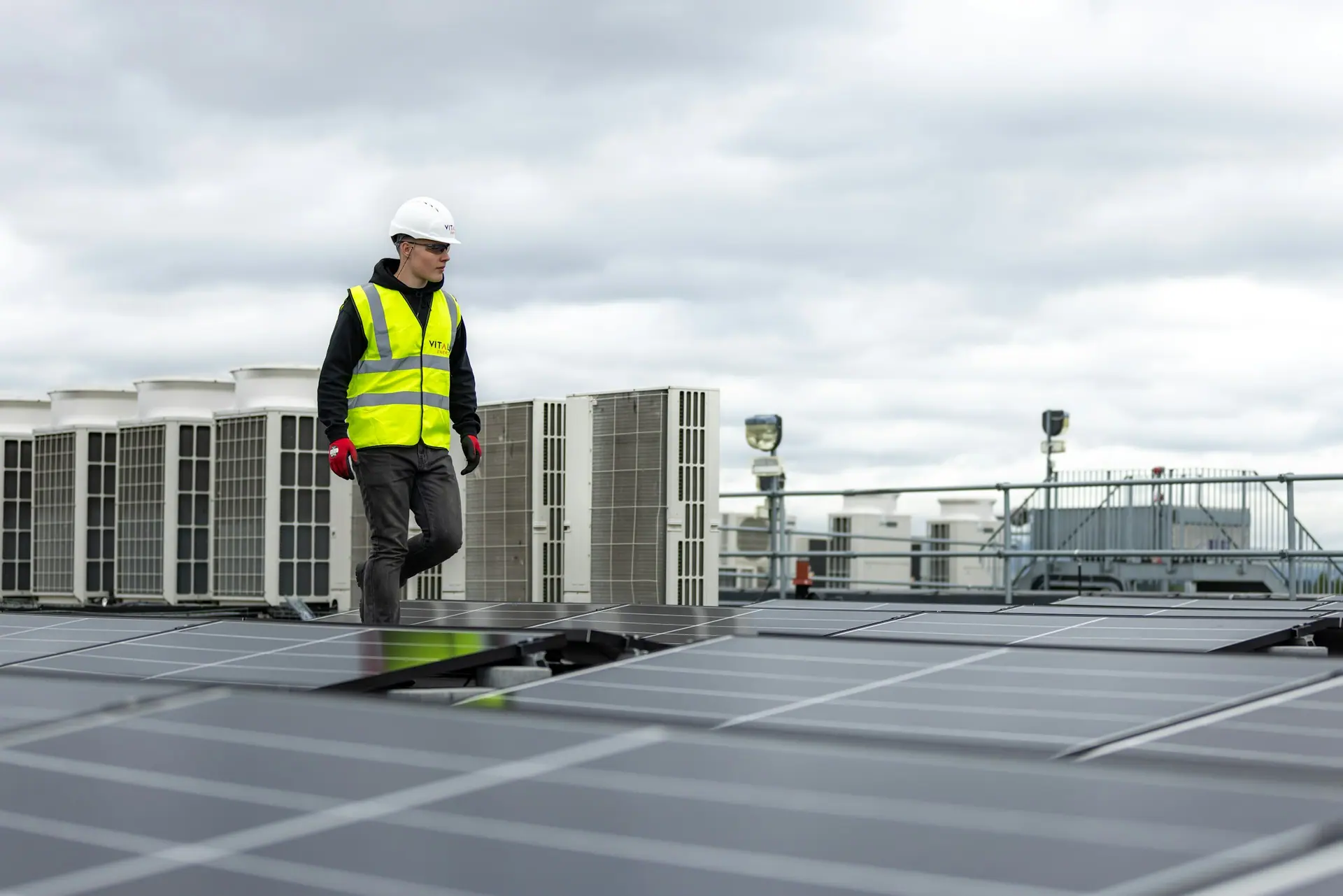 Technician in a hard hat and hi-vis vest walking across a rooftop beside condenser units